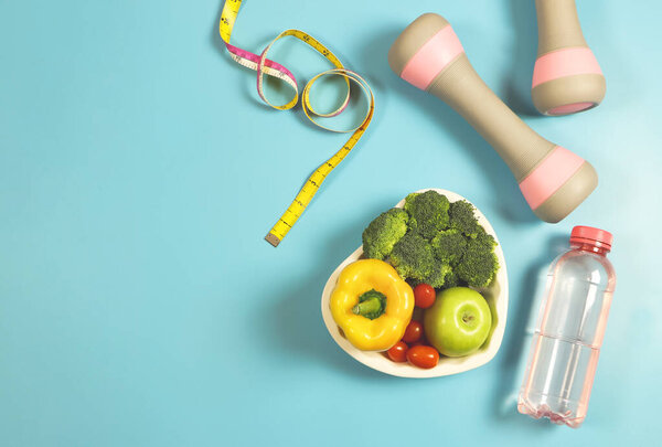 top view of vegetables  capsicum, broccoli, tomatoes and apple  in heart shape plate  on blue  background with pink bottle of water, gray pink dumbbells and measure tape. Healthy lifestyle and weight loss.