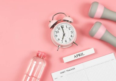 Üst ya da düz alışkanlık takip kitabı, ahşap takvim Nisan, gri pembe dambıllar, bir şişe su ve pembe arkaplanlı pembe çalar saat. Kişisel gelişim ve aktif yaşam tarzı.