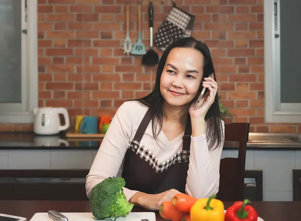 亚洲家庭主妇在厨房用手机手持装置准备食物的肖像 一个女人坐在厨房里 拿着蔬菜 用手机聊天 心情很好 多重任务 — 图库照片