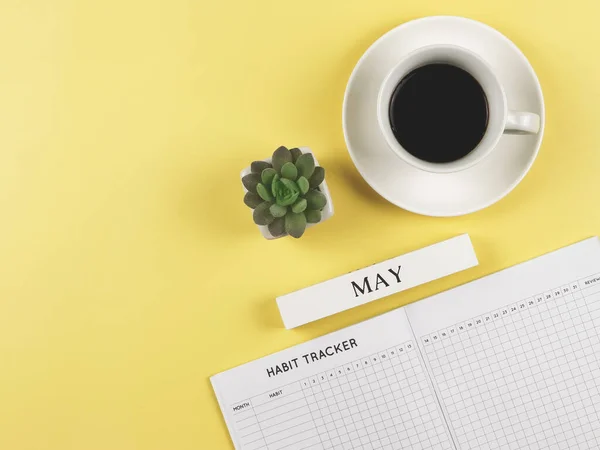 Top view or flat lay of habit tracker book, wooden calendar May, a cup of black coffee and succulent plant pot on yellow background with copy space.