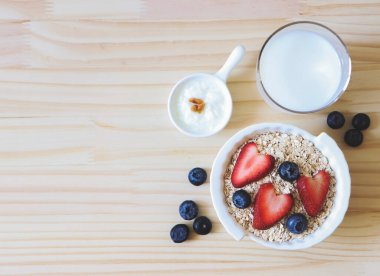 Beyaz kâsede yulaf ya da tahıl gevreği, taze yaban mersini, çilek, bir bardak süt ve yoğurt ve ahşap masada. Sağlıklı kahvaltı kavramı.