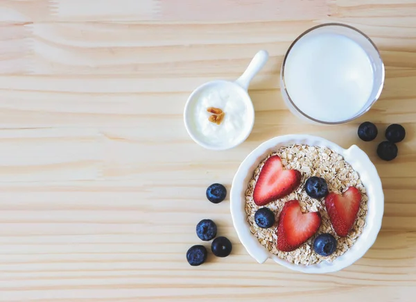 Beyaz kâsede yulaf ya da tahıl gevreği, taze yaban mersini, çilek, bir bardak süt ve yoğurt ve ahşap masada. Sağlıklı kahvaltı kavramı.