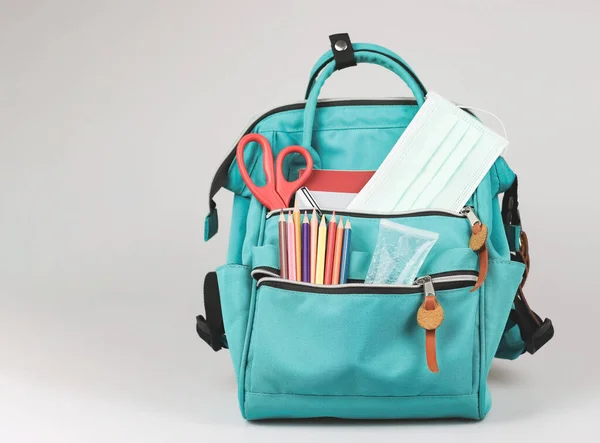 Front view  of  backpack with school supplies , medical mask and alcohol sanitizer gel on white  background with copy space. COVID-19 prevention while going back  to school  and new normal  concept.