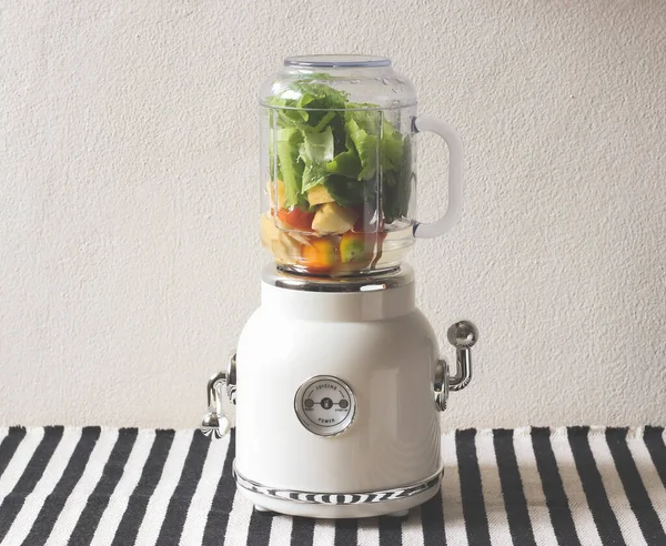 Front view of  white vintage blender or smoothie maker withe vegetables, tomatos, apples and water   on  black and white stripe table cloth and white wall.