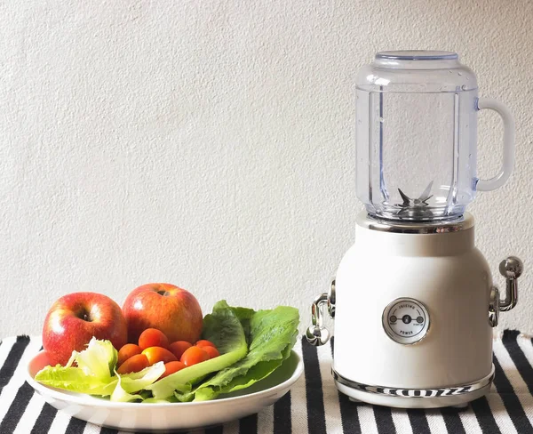 Front view of empty white vintage blender or smoothie maker  with a plate of vegetables, tomatoes and apples on  black and white stripe table cloth and white wall. Preparing for smoothie making.