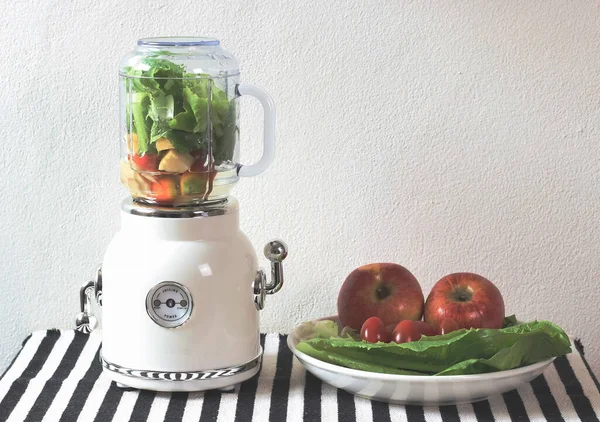 Front view of  white vintage blender or smoothie maker  with a plate of vegetables, tomatoes and apples on  black and white stripe table cloth and white wall. Healthy drink making.