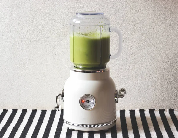 Front view of  white vintage blender or smoothie maker machine  working on making green smoothie  on  black and white stripe table cloth and white wall. Healthy drink making.