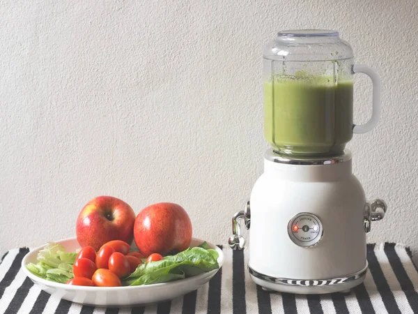 Front view of  white vintage blender or smoothie maker  with a plate of vegetables, tomatoes and apples on  black and white stripe table cloth and white wall. Healthy drink making.