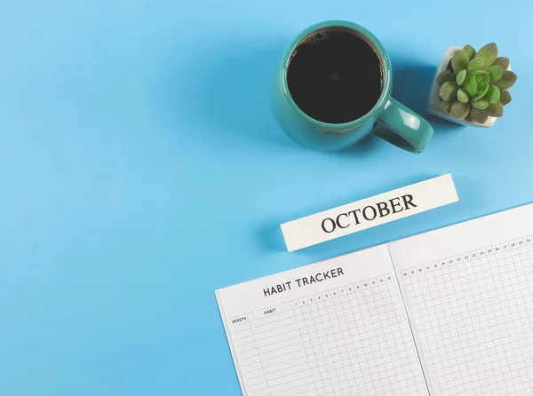 Top view or flat lay of habit tracker book, wooden calendar October,  blue cup of black coffee and succulent plant pot on blue background with copy space.