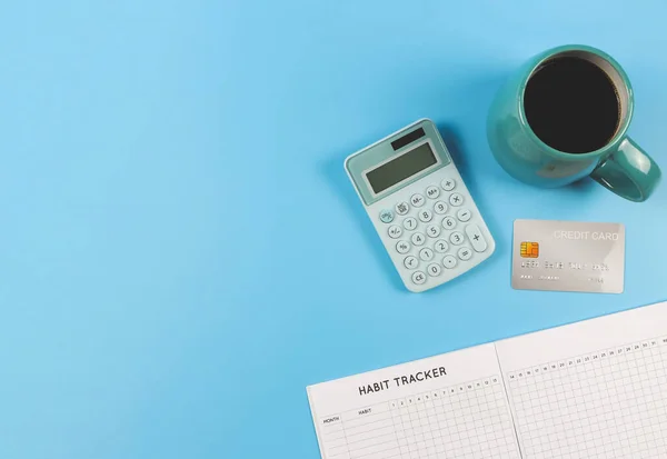 Top view or flat lay of habit tracker book,  blue cup of black coffee, blue calculator and credit card on blue background with copy space. Business and good  financial habit