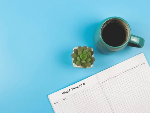 Top view or flat lay of habit tracker book with  blue  cup of black coffee and succulent plant pot on blue background with copy space.