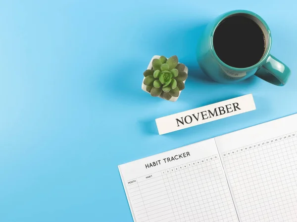 Top view or flat lay of habit tracker book with  wooden calendar november, blue  cup of black coffee and succulent plant pot on blue background with copy space.