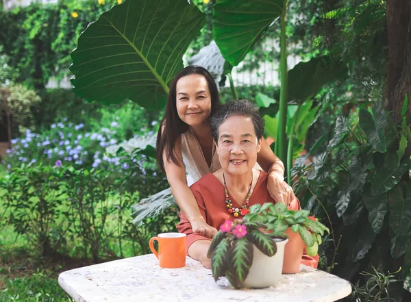 Portrait of Asian senior woman sitting with her daughter  at white table in beautiful garden, daughter hugging her mother from the back,  smiling and looking at camera.