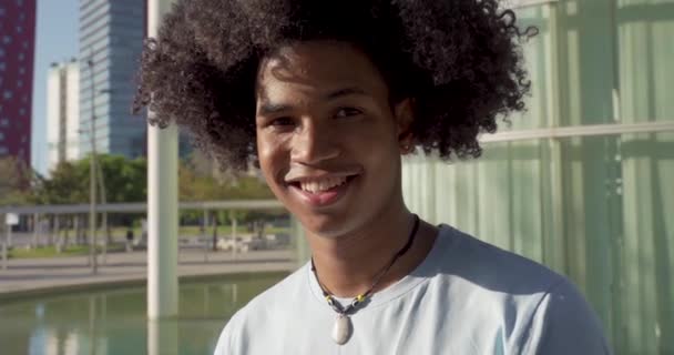 Νεαρός όμορφος μαύρος Αφροαμερικάνος κοιτάζει την κάμερα χαμογελώντας στην πανεπιστημιούπολη σε μια ηλιόλουστη μέρα σε αργή κίνηση. — Αρχείο Βίντεο