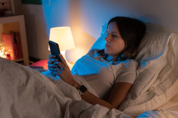 Belle jeune fille regardant son téléphone sur le lit au milieu de la nuit. Concept de technologie au lit. — Photo