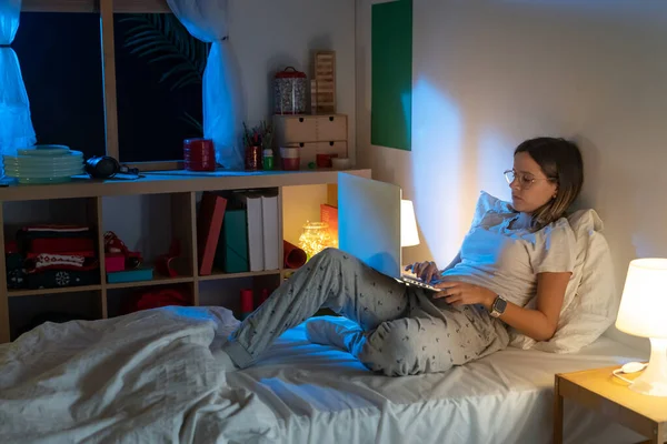 Belle jeune fille assise sur le lit la nuit travaillant avec son ordinateur portable au milieu de la nuit. — Photo