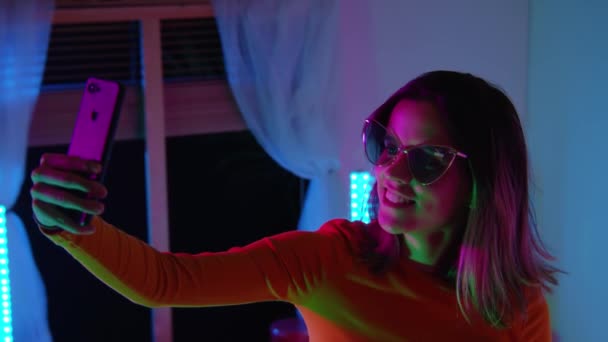 Hermosa chica joven tomando selfie con su teléfono en la cama con sala de colores de neón. Tecnología en la cama concepto. — Vídeos de Stock