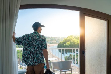 Otel odasında bavulu pencerenin yanında duran bir turist. Seyahat ve tatil kavramı.