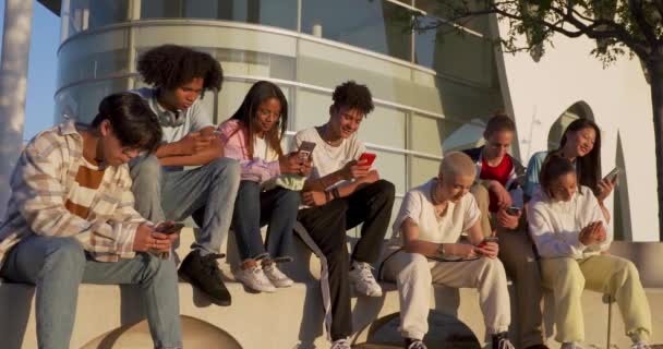 Grande grupo de adolescentes felizes olhando seus telefones inteligentes em um banco na rua do campus estudantil. — Vídeo de Stock