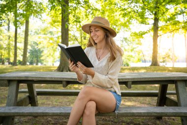 Yeşil Park 'ta gülümseyen ve kitap okuyan genç bir kadın. Doğa, konsept hobi ve yaşam tarzı üzerine roman. İnanç, maneviyat ve din. Sevimli kız öğrencinin açık hava fotoğrafı. Yüksek kalite