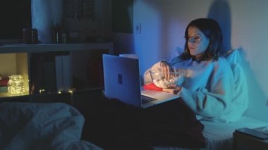 Genç kız geceleri dizüstü bilgisayarıyla film izleyip patlamış mısır yiyor. Yavaş çekim kırmızı kamera 4K. Yüksek kalite 4k görüntü