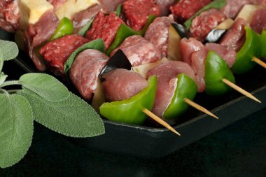 Macro close up of raw meat skewers before cooking on a dark background with sage leaves. 