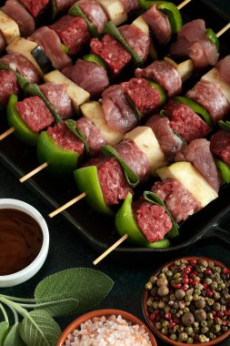 Macro close up of raw meat skewers before cooking on a dark background with sage leaves and mixed pepper. 