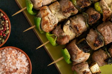 Macro close up of baked meat skewers on a green dish with sage leaves, salt and mixed pepper. 