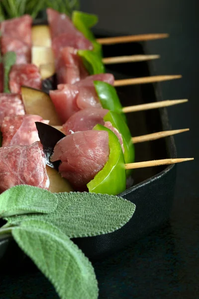 Macro close up of raw meat skewers before cooking on a dark background with sage leaves. 