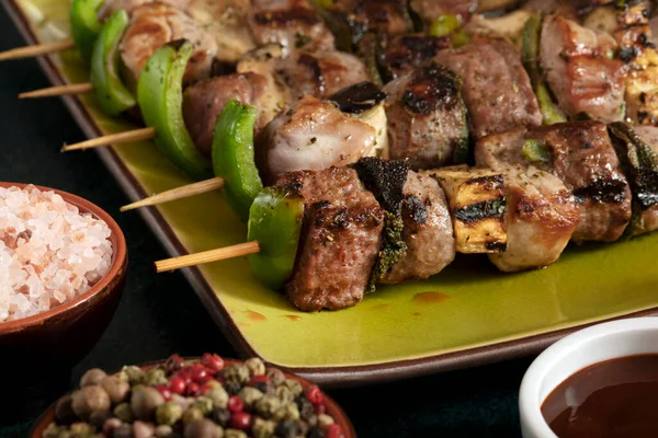 Macro close up of baked meat skewers on a green dish with sage leaves, salt and mixed pepper. 
