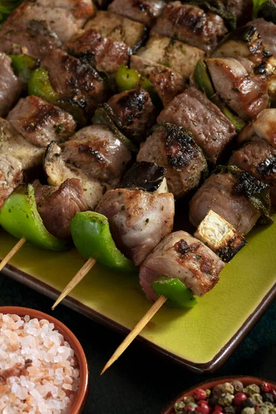 Macro close up of baked meat skewers on a green dish with sage leaves, salt and mixed pepper. 