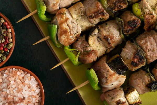 Macro close up of baked meat skewers on a green dish with sage leaves, salt and mixed pepper. 