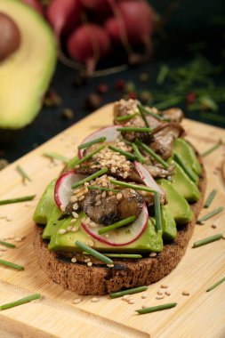 An avocado toast with mushrooms, radish, sesame seeds and chive on a wooden cutting board. 