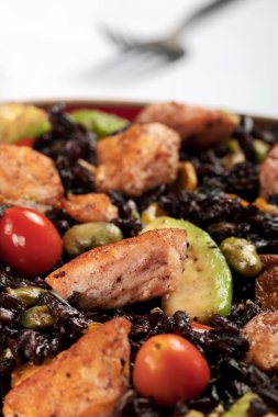A red bowl of black rice with baked salmon, avocado, tomatoes, chili peppers and edamame. 