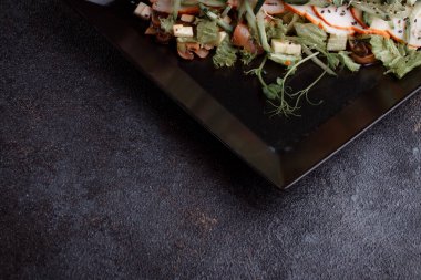 Summer salad with pieces of bacon, fresh cucumbers, cheese cubes and mushrooms. Green salad on a black plate on a dark background. Free space for an inscription. View from above.