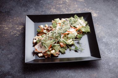 Summer salad with pieces of bacon, fresh cucumbers, cheese cubes and mushrooms. Green salad on a black plate on a dark background. Free space for an inscription. View from above.
