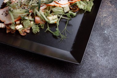 Summer salad with pieces of bacon, fresh cucumbers, cheese cubes and mushrooms. Green salad on a black plate on a dark background. Free space for an inscription. View from above.