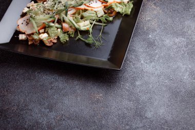 Summer salad with pieces of bacon, fresh cucumbers, cheese cubes and mushrooms. Green salad on a black plate on a dark background. Free space for an inscription. View from above.