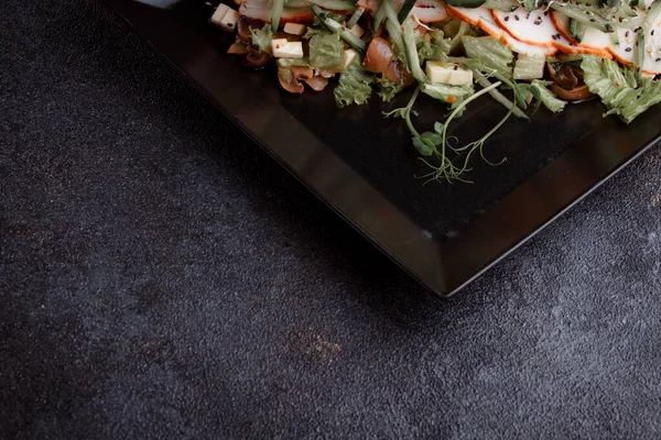 Summer salad with pieces of bacon, fresh cucumbers, cheese cubes and mushrooms. Green salad on a black plate on a dark background. Free space for an inscription. View from above.