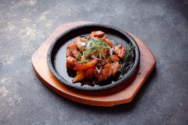 Hot frying pan with pieces of fried meat in caramelized sauce. Cooked and fresh vegetables as garnish and garnish.