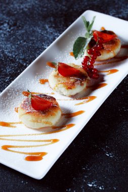 Çilek ve üzümle süslenmiş süzme peynir krepleri. Koyu mavi arka planda güzel bir yemek servisi. Restoran tatlı menüsü.