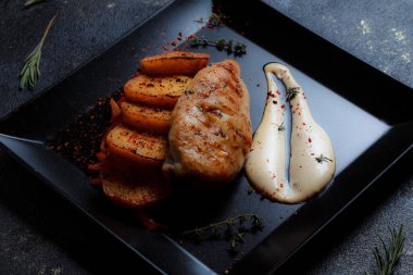 Baharatlı ızgara tavuk bifteği ve siyah bir tabakta fırında patates fotokopi alanı olan koyu dokulu bir arka planda..