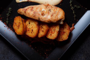 Baharatlı ızgara tavuk bifteği ve siyah bir tabakta fırında patates fotokopi alanı olan koyu dokulu bir arka planda..
