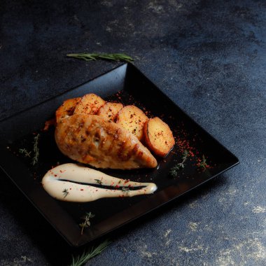 Baharatlı ızgara tavuk bifteği ve siyah bir tabakta fırında patates fotokopi alanı olan koyu dokulu bir arka planda..