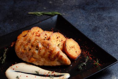 Grilled chicken steak with spices and baked potatoes on a black plate on a dark textured background with copy space.
