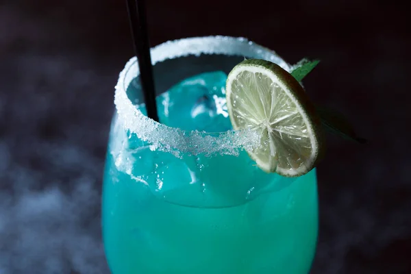 Alcoholic cocktail blue lagoon. Green cocktail drink with lime on a dark blue textured background.