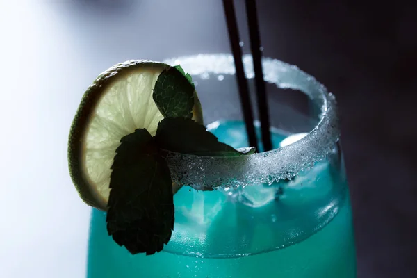 Alcoholic cocktail blue lagoon. Green cocktail drink with lime on a dark blue textured background.