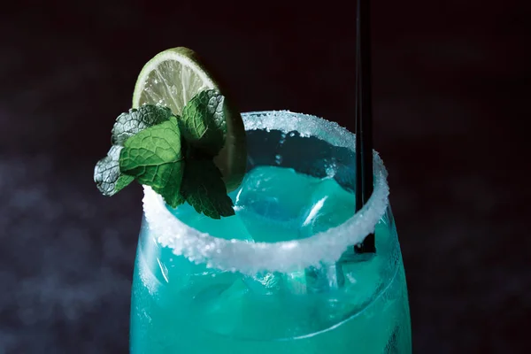 Alcoholic cocktail blue lagoon. Green cocktail drink with lime on a dark blue textured background.