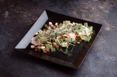 Summer salad with pieces of bacon, fresh cucumbers, cheese cubes and mushrooms. Green salad on a black plate on a dark background. Free space for an inscription. View from above.