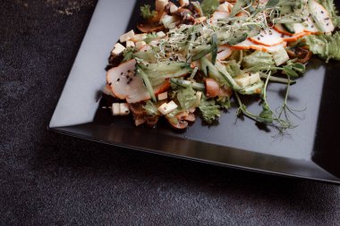 Summer salad with pieces of bacon, fresh cucumbers, cheese cubes and mushrooms. Green salad on a black plate on a dark background. Free space for an inscription. View from above.